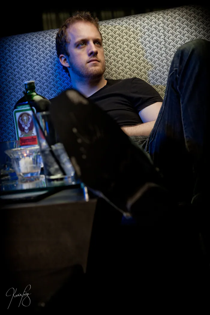 Low-key lounge portrait — man in black seated beside a bottle-lined table