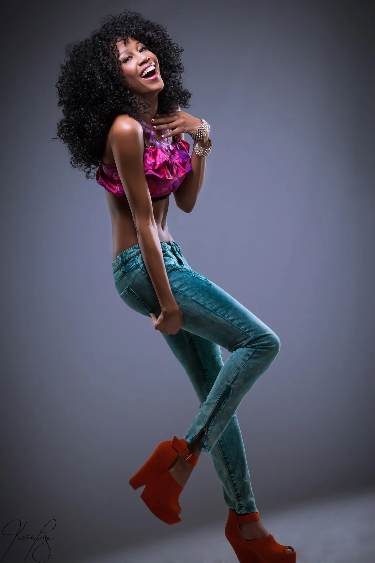 Studio fashion shot — model in teal jeans and pink top holding a red heel